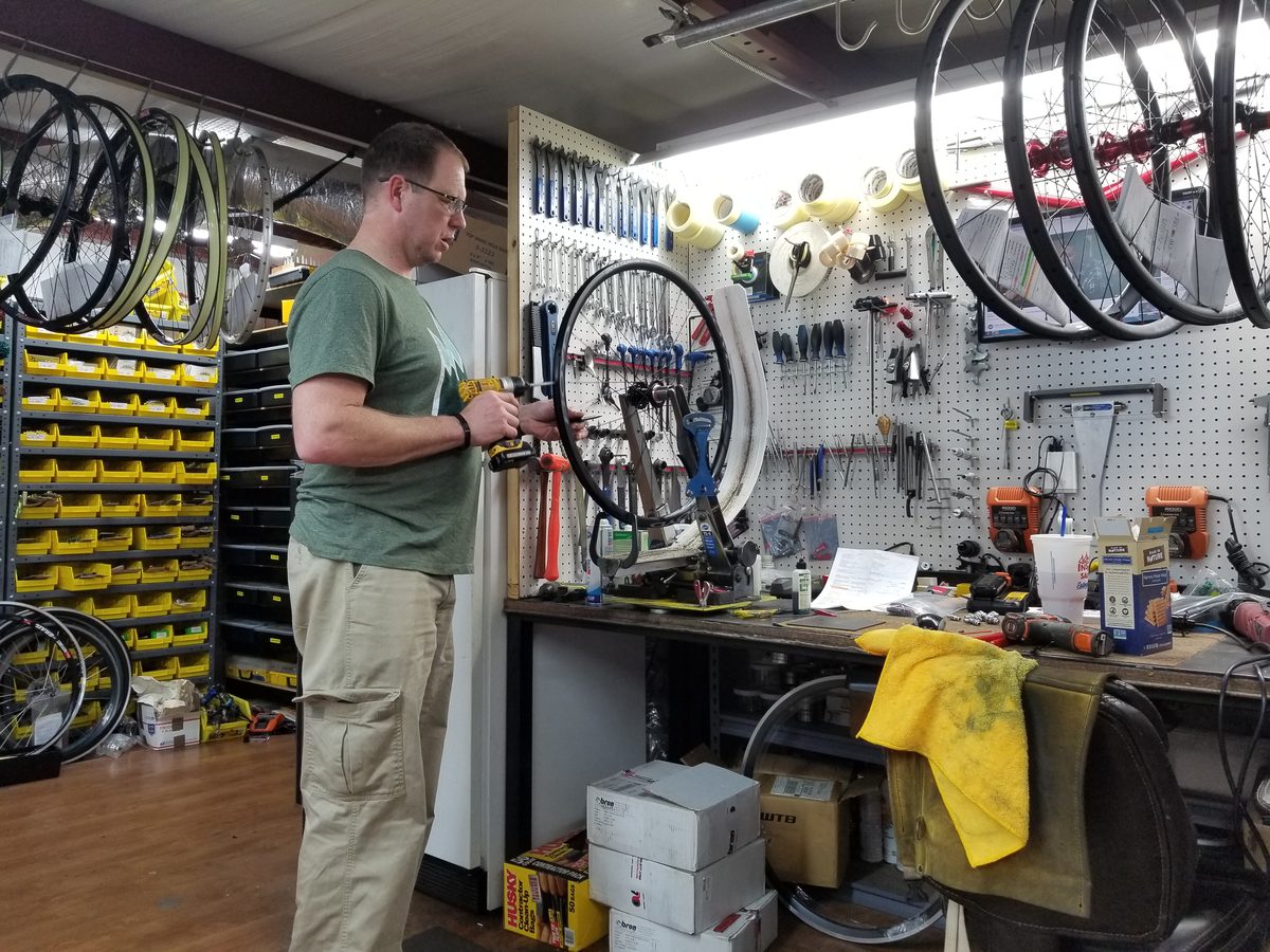 A wheelbuilder truing a wheel at the bench, with finished wheels and build sheets hanging above